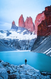 man standing on rock facing body of water and mountain