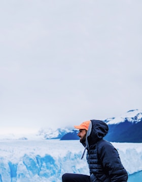 A man wearing a dark jacket and an orange cap kneels in front of a large ice glacier. Snow-covered mountains can be seen in the background against an overcast sky.