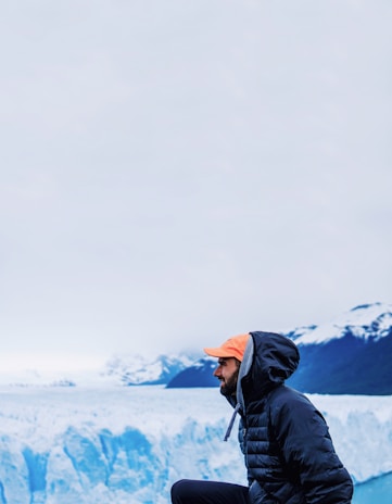 A man wearing a dark jacket and an orange cap kneels in front of a large ice glacier. Snow-covered mountains can be seen in the background against an overcast sky.