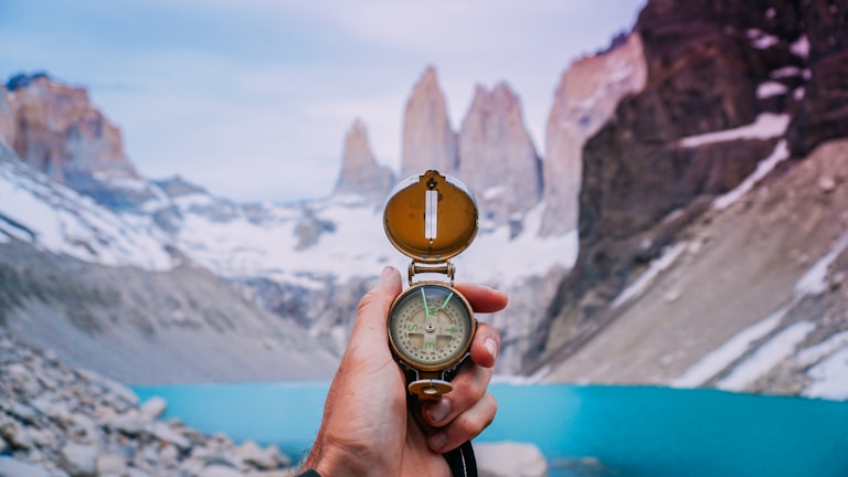A hand holding a compass, pointing towards a stunning mountainous landscape. Snow-capped peaks rise dramatically in the background, towering over a tranquil turquoise lake. The scene conveys a sense of adventure and exploration.