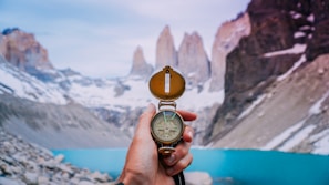 A hand holding a compass, pointing towards a stunning mountainous landscape. Snow-capped peaks rise dramatically in the background, towering over a tranquil turquoise lake. The scene conveys a sense of adventure and exploration.