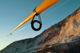 A close-up view of a fishing rod with a focus on the guide ring. The rod is angled diagonally across the image with a clear blue sky in the background and a rocky cliffside on the right.