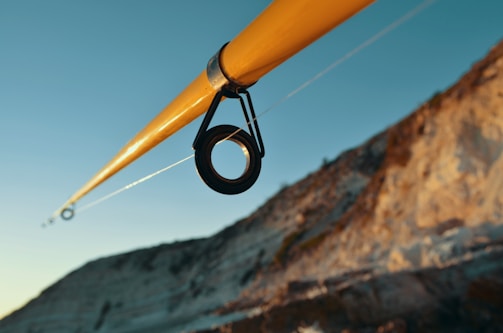 A close-up view of a fishing rod with a focus on the guide ring. The rod is angled diagonally across the image with a clear blue sky in the background and a rocky cliffside on the right.