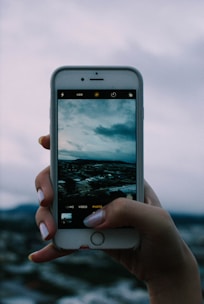 A hand holding a smartphone displays an image of a cloudy sky and a landscape scene in the camera app. The phone screen contrasts with the blurred, cloudy background, suggesting the phone is capturing a similar view.