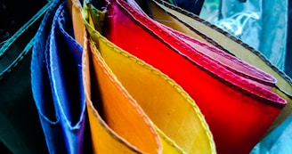 A collection of vibrantly colored fabric pieces arranged in a fan-like pattern. The fabrics are in various shades including blue, purple, red, orange, and yellow. The textures and stitches of the fabrics are clearly visible, creating a rich visual display of colors and materials.