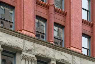 A section of a building facade featuring a combination of red brick and stone elements. The red brick section includes ornate columns positioned between rectangular windows. Below the windows, stone detailing with decorative carvings enhances the architectural design.