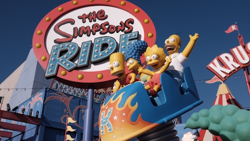 A vibrant amusement park ride featuring cartoon characters in a roller coaster cart beneath a large, colorful sign. The characters appear animated and joyful, with exaggerated facial expressions. The surroundings include bright, playful architecture and additional amusement park elements.