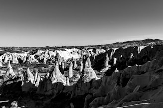 A breathtaking landscape featuring a series of rugged, pointed rock formations in a rocky desert area. The geological structures resemble a surreal and otherworldly environment, possibly with deep shadows creating dramatic contrasts. The terrain appears weathered, adding a timeless quality to the scene.