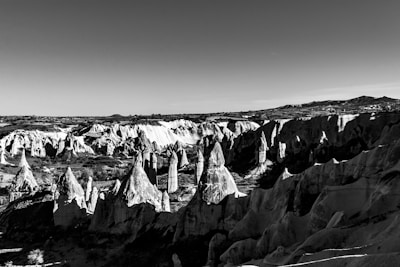 A breathtaking landscape featuring a series of rugged, pointed rock formations in a rocky desert area. The geological structures resemble a surreal and otherworldly environment, possibly with deep shadows creating dramatic contrasts. The terrain appears weathered, adding a timeless quality to the scene.