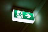 A green emergency exit sign with a pictogram of a person running and an arrow pointing to the right. The sign is illuminated and positioned on a ceiling, with a slight shadow cast on the wall.