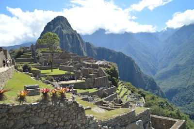 Ancient stone structures and terraces are set against a backdrop of lush green mountains and blue skies with scattered clouds. The site is bustling with people exploring the historical ruins, likely a famous archaeological landmark.