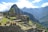 Ancient stone structures and terraces are set against a backdrop of lush green mountains and blue skies with scattered clouds. The site is bustling with people exploring the historical ruins, likely a famous archaeological landmark.