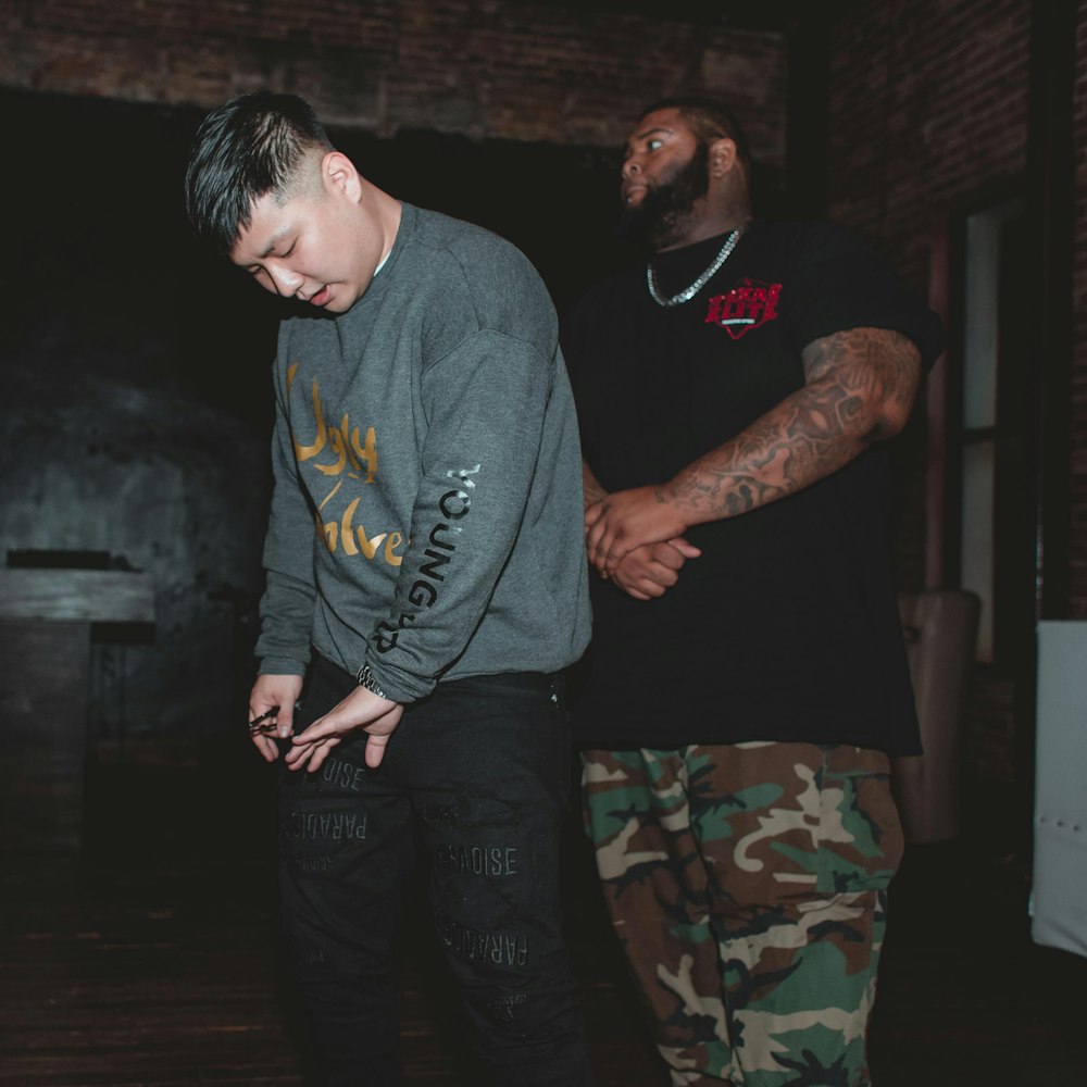 man wearing black t-shirt and camouflage cargo pants standing behind man in gray sweatshirt and black pants