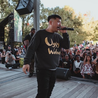 A person is performing on stage with a microphone in front of an engaged crowd. The performer is wearing a dark sweater with the words 'Ugly Wolves' in gold lettering. The audience appears attentive and diverse, with some people holding up their phones to capture the moment. The backdrop shows trees and a clear sky, indicating an outdoor setting.