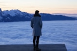 A person stands on a platform overlooking a vast expanse of clouds with snow-capped mountains in the background. The sky has a soft gradient, transitioning from light pink to a deeper blue, suggesting either sunrise or sunset. The person is wearing a long coat and has their hands in their pockets.