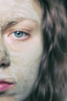 A close-up of a person's face featuring an earthy, cracked clay facial mask covering their skin. The focus is on a single, striking blue eye, with natural light highlighting the texture of the mask. Long brown hair frames the side of the face, adding contrast to the pale clay color.