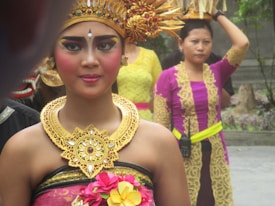 A woman adorned in traditional attire featuring intricate gold headgear and jewelry stands in focus. Her makeup is bold and colorful, emphasizing her eyes. Behind her, another woman is visible wearing a vibrant purple and yellow outfit, holding an object on her head. The background includes more people and stone structures, suggesting a cultural event or ceremony.