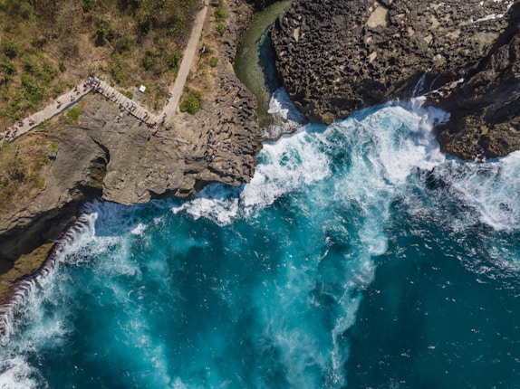 An aerial view captures a rugged coastline with vibrant turquoise waves crashing against jagged rocks. A pathway winds along the cliff, where small groups of people gather, overlooking the ocean. Vegetation grows sparsely on the rocky terrain.
