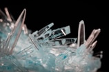 A cluster of sharp, translucent blue crystals intertwined with clear, elongated spikes against a dark background. The crystals have a textured and reflective surface, catching and diffusing light to create a shimmering effect.