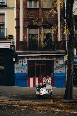 A vintage scooter is parked in front of a garage named 'Talleres de Motos', with a storefront featuring red and white stripes under a brick residential building. The street is lined with fallen leaves, suggesting autumn. The building's facade has a mix of brick and plaster with a small balcony, and signs indicating motorbike repair services.