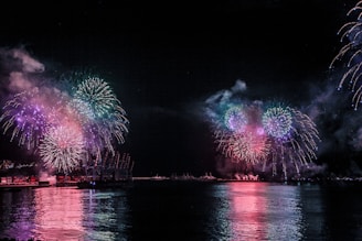 Vibrant fireworks illuminate the night sky over a body of water, reflecting colorful explosions on the surface. Smoke lingers in the air, creating a festive and lively atmosphere. The scenery captures both the waterfront and distant structures silhouetted against the bursts of color.