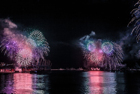 Vibrant fireworks illuminate the night sky over a body of water, reflecting colorful explosions on the surface. Smoke lingers in the air, creating a festive and lively atmosphere. The scenery captures both the waterfront and distant structures silhouetted against the bursts of color.