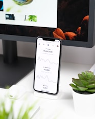 A smartphone displays analytics data with graphs showing views, downloads, and likes. It is resting vertically on a white dock next to a small potted succulent plant. In the background, a large computer monitor is partially visible, along with a closed laptop and a white wireless earbud case.