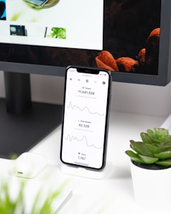 A smartphone displays analytics data with graphs showing views, downloads, and likes. It is resting vertically on a white dock next to a small potted succulent plant. In the background, a large computer monitor is partially visible, along with a closed laptop and a white wireless earbud case.