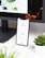 A smartphone displays analytics data with graphs showing views, downloads, and likes. It is resting vertically on a white dock next to a small potted succulent plant. In the background, a large computer monitor is partially visible, along with a closed laptop and a white wireless earbud case.