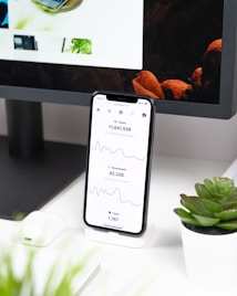 A smartphone displays analytics data with graphs showing views, downloads, and likes. It is resting vertically on a white dock next to a small potted succulent plant. In the background, a large computer monitor is partially visible, along with a closed laptop and a white wireless earbud case.