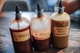 Three sauce bottles are placed on a wooden surface. Each bottle has a handwritten label: 'Spicy!', 'House', and 'Sweet & Tangy Mustard'. The bottles have squeeze tops and contain different colored sauces.