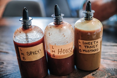 Three sauce bottles are placed on a wooden surface. Each bottle has a handwritten label: 'Spicy!', 'House', and 'Sweet & Tangy Mustard'. The bottles have squeeze tops and contain different colored sauces.