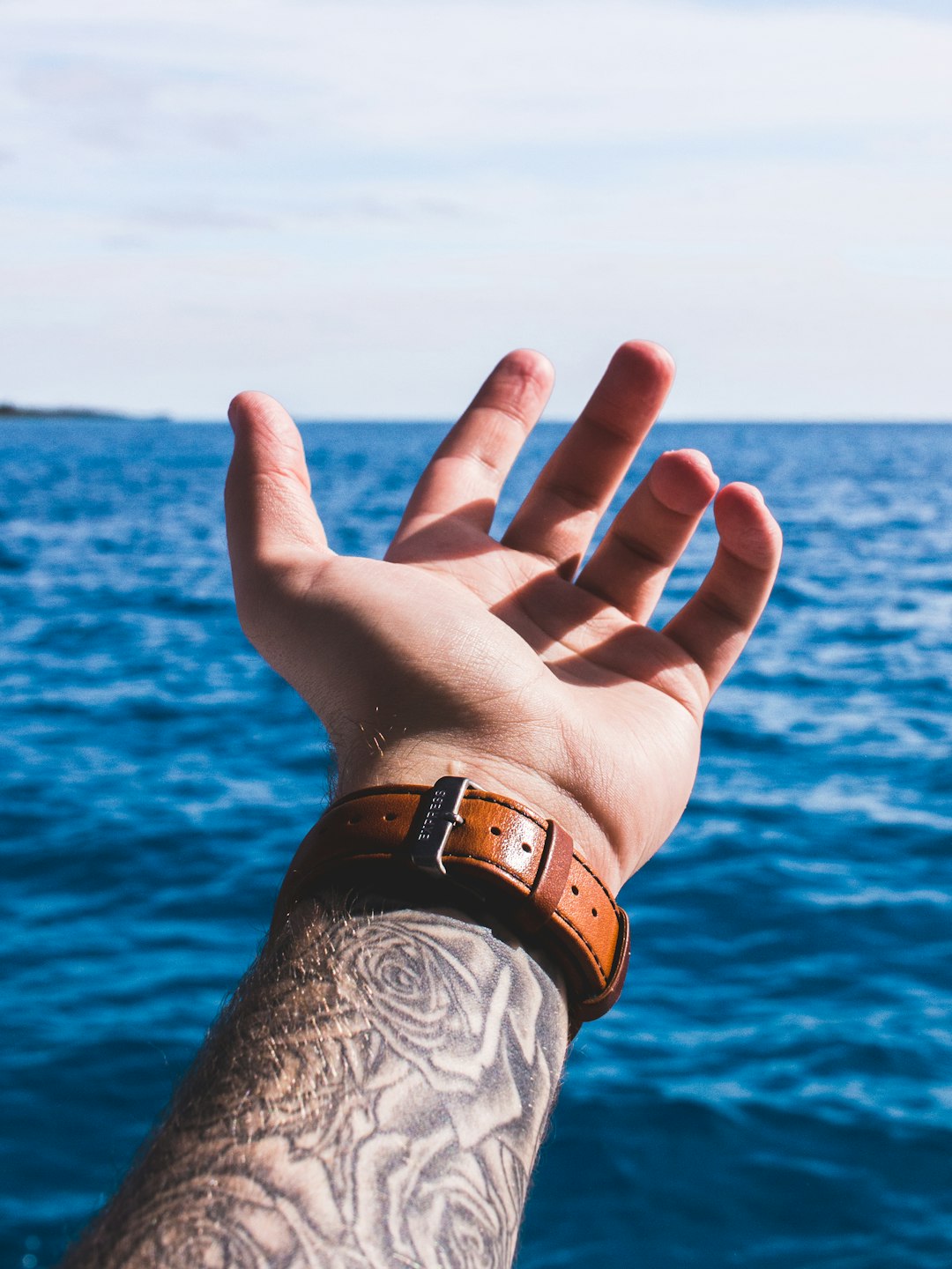 person with arm sleeved tattoo wearing brown leather strap watch holding out hand above water