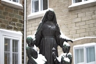 A bronze statue depicting a figure in a hooded robe, likely representing a nun, surrounded by two children. The statue is covered with patches of snow, and it stands in front of a building with stone walls and windows.