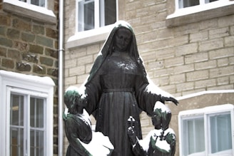A bronze statue depicting a figure in a hooded robe, likely representing a nun, surrounded by two children. The statue is covered with patches of snow, and it stands in front of a building with stone walls and windows.