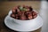 A white bowl filled with dark, reddish-brown chips set on a white plate. A small green garnish is placed atop the chips. The background features a blurred brown surface, enhancing the focus on the dish.