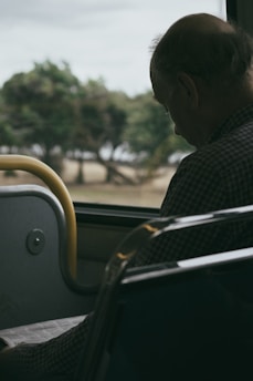 An elderly man with a balding head sits on a bus, gazing downward. He appears to be holding or reading something on his lap. The background reveals a blurred view of trees and a grassy field outside the bus window, suggesting movement.