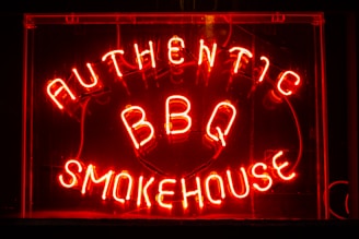 Bright neon letters spell out 'Authentic BBQ Smokehouse' in a vivid red color on a dark background. The signage is enclosed in a glass frame and illuminated, creating a warm, inviting glow.