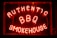 Bright neon letters spell out 'Authentic BBQ Smokehouse' in a vivid red color on a dark background. The signage is enclosed in a glass frame and illuminated, creating a warm, inviting glow.