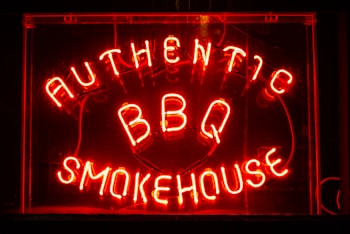 Bright neon letters spell out 'Authentic BBQ Smokehouse' in a vivid red color on a dark background. The signage is enclosed in a glass frame and illuminated, creating a warm, inviting glow.