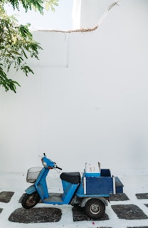 A blue scooter with a rear storage box is parked on a cobblestone path against a white wall. Overhead, branches of a tree provide a touch of greenery.