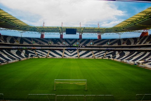 A large, empty football stadium with distinctive black and white patterned seating. The field is covered with well-maintained green grass, and a single goal post is visible at one end. The stadium has a partially covered roof with a modern design featuring red structural elements.