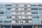 A multi-story residential building with a symmetrical facade, featuring rows of windows and balconies. The glass windows are framed in white and there are several plants on the balconies. The overall structure appears modern with an urban atmosphere.