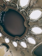 An ornate ceiling featuring intricate plasterwork and decorative elements, with multiple oval-shaped light fixtures. The design includes floral motifs and swirling patterns, creating an elegant and classical appearance.