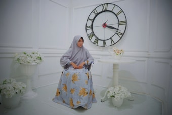 A woman in a light gray hijab sits in a room with white walls. She wears a long dress with a pattern of yellow leaves. Behind her is a large wall clock with black frames and Roman numerals. There are white floral arrangements in vases surrounding her.