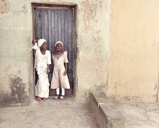 Two children dressed in light-colored traditional attire stand in a doorway. The worn-out wall behind them is textured and mildly discolored, suggesting an older structure. Their expressions are neutral to curious.