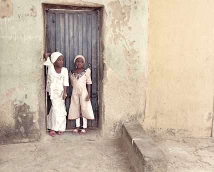 Two children dressed in light-colored traditional attire stand in a doorway. The worn-out wall behind them is textured and mildly discolored, suggesting an older structure. Their expressions are neutral to curious.