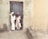 Two children dressed in light-colored traditional attire stand in a doorway. The worn-out wall behind them is textured and mildly discolored, suggesting an older structure. Their expressions are neutral to curious.