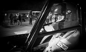 A person wearing a racing helmet and suit sits inside a vehicle with a visible roll cage. In the background, there are a few people standing, and the scene is illuminated by artificial light, likely at a racetrack or similar setting.