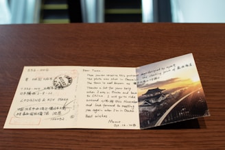 A handwritten postcard displayed on a wooden table, featuring a photograph of a traditional Japanese building at sunset. The postcard includes text in English and Chinese/Japanese with postmarks and addresses. The photograph shows a scenic view of a structure with curved roofs and a picturesque sunset.